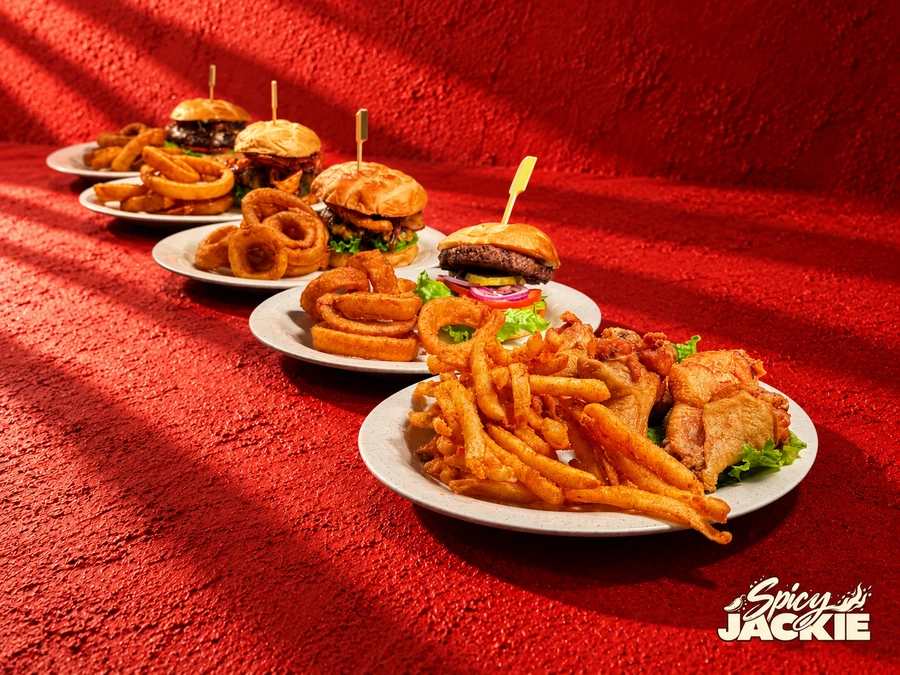 A diagonal lineup of burgers with onion rings and a sandwich with fries on white plates against a textured red background