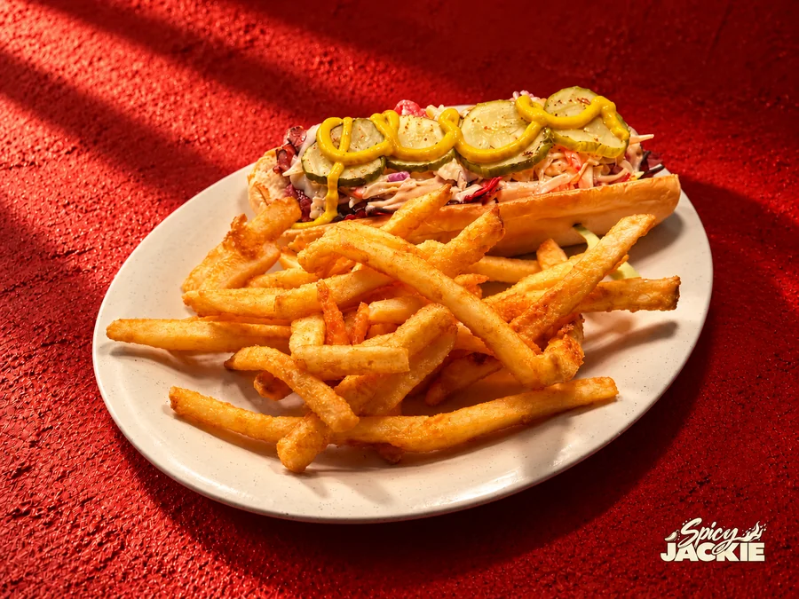 A meat sandwich with slaw, pickle, and mustard beside fries on a white plate
