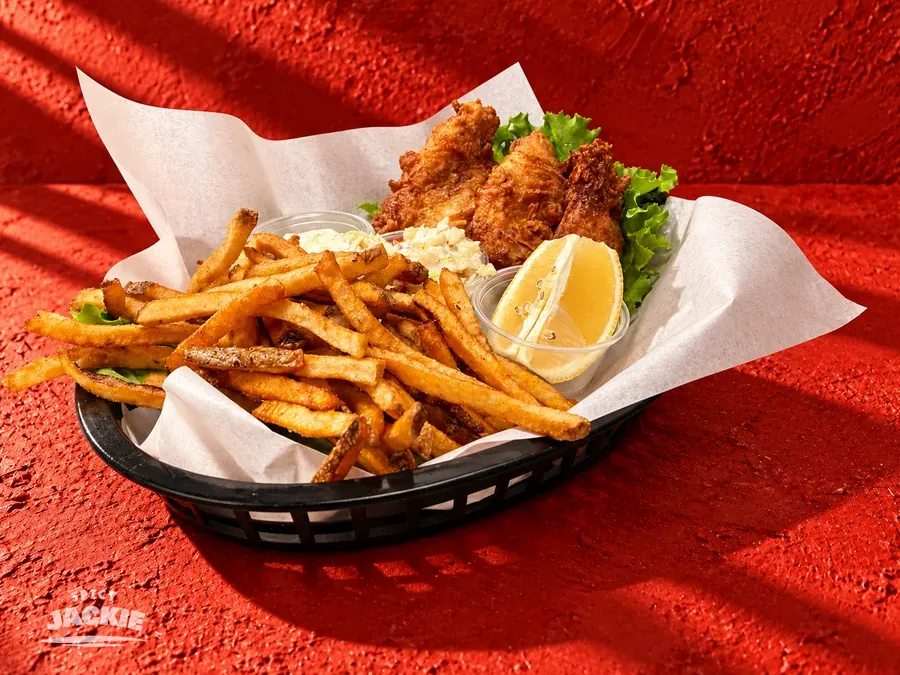 Basket of battered fish and fries with a lemon wedge and small side cups