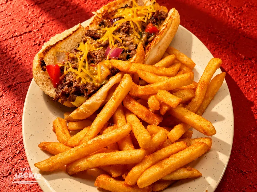 Filled sandwich in a long roll topped with shredded cheese beside fries on a white plate