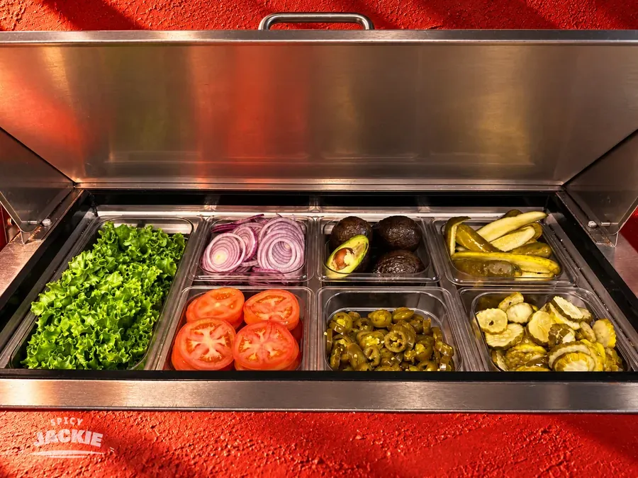An open refrigerated ingredient station filled with lettuce, onion, tomato, jalapeños, pickles, and avocados