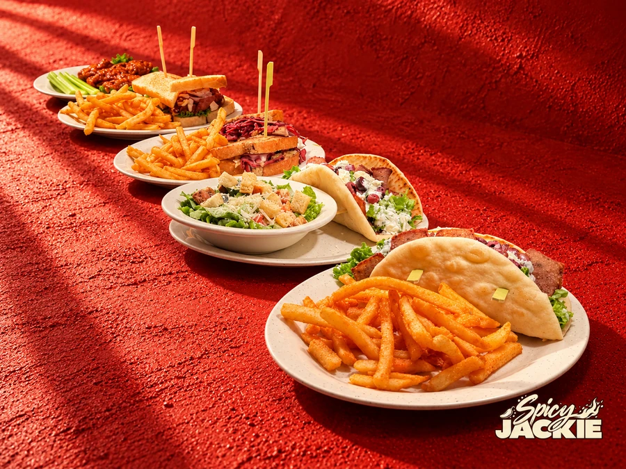 A diagonal lineup of pita sandwiches, salad, toasted sandwiches, fries, and wings with celery on a textured red surface
