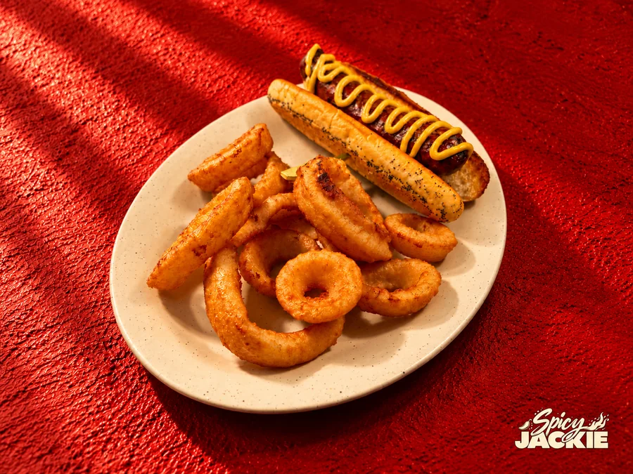 Grilled sausage with mustard in a poppy-seed roll beside golden onion rings on a white plate