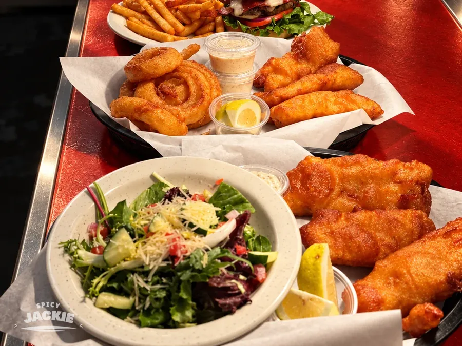 Multiple restaurant plates lined up along the kitchen counter, including salad, fried fish, burgers, fries, and onion rings